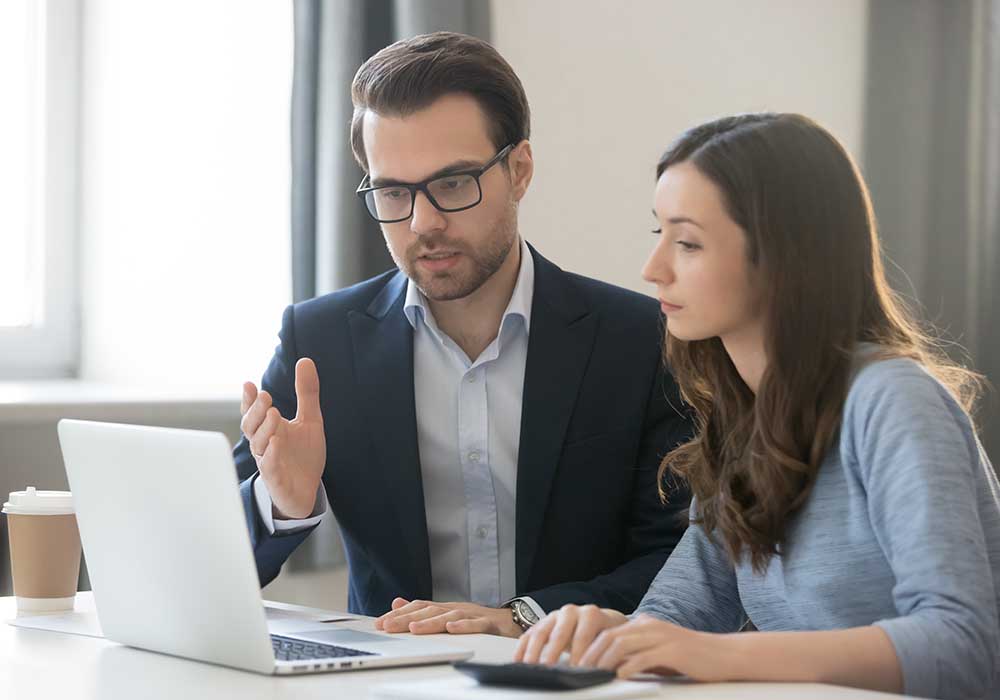 Geschäftsmann erklärt jüngerer Mitarbeiterin etwas an ihrem Laptop (PowerPoint-Dienstleister)